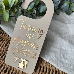 Puede incluir: Un colgador de puerta beige con letras doradas que dice "Tanning in progress PLEASE DO NOT DISTURB". El colgador tiene una parte superior curva para colgar y presenta iconos dorados de una pistola de bronceado en aerosol y una cabina de bronceado.