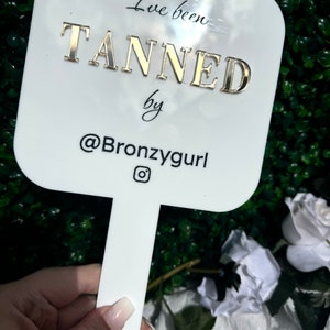 May include: White handheld mirror with a gold inscription that reads "I've been TANNED by @Bronzygurl" with an Instagram icon.