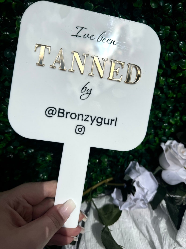 May include: White handheld mirror with a gold inscription that reads "I've been TANNED by @Bronzygurl" with an Instagram icon.