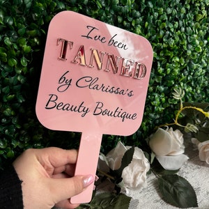 May include: Pink hand-held fan with the text "I've been TANNED by Clarissa's Beauty Boutique" in black lettering with gold glitter letters.