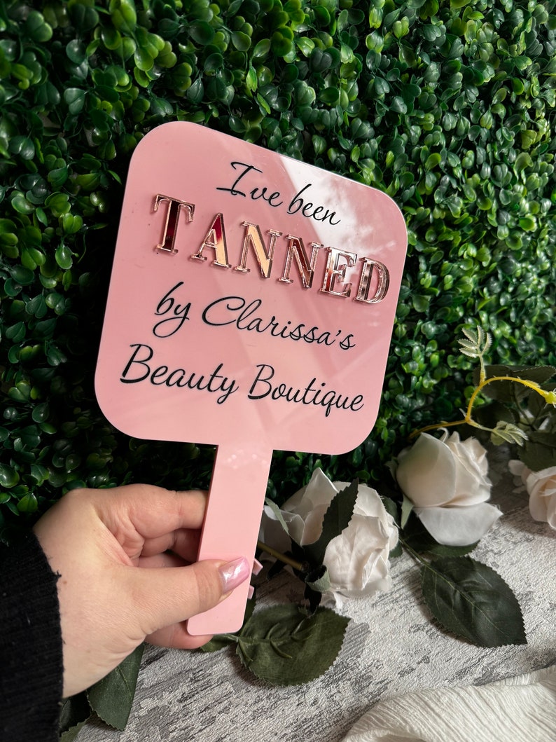 May include: Pink hand-held fan with the text "I've been TANNED by Clarissa's Beauty Boutique" in black lettering with gold glitter letters.