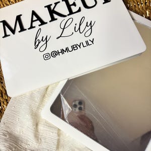 K&ouml;nnte beinhalten: Ein wei&szlig;es rechteckiges Schild mit dem schwarzen Text MAKEUP by Lily und dem Instagram-Handle @HMUBYLILY. Darunter befindet sich ein rechteckiger Spiegel mit einer reflektierenden Oberfl&auml;che, die teilweise mit einer Schutzfolie bedeckt ist.
