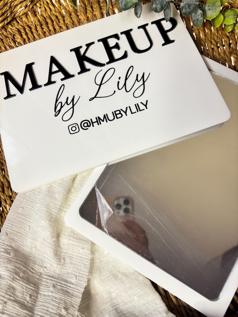 K&ouml;nnte beinhalten: Ein wei&szlig;es rechteckiges Schild mit dem schwarzen Text MAKEUP by Lily und dem Instagram-Handle @HMUBYLILY. Darunter befindet sich ein rechteckiger Spiegel mit einer reflektierenden Oberfl&auml;che, die teilweise mit einer Schutzfolie bedeckt ist.
