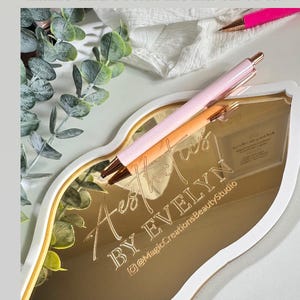 May include: A gold-colored, lip-shaped tray with a white border. The tray has the words "Aest" and "BY EVELYN" engraved on it, along with the social media handle "@MagicCreationsBeautyStudio".