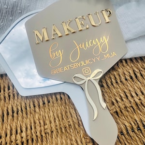 May include: A hand-held makeup mirror with a light gray handle and mirror frame. The mirror's face is a light gray color with the words "MAKEUP by Juicyy" in gold lettering. A decorative bow is attached to the handle. The Instagram handle @BEATSBYJUICYY_MUA is also visible.