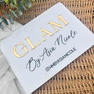 K&ouml;nnte beinhalten: Ein rechteckiges wei&szlig;es Acrylschild mit der goldenen erhabenen Aufschrift GLAM, dem schwarzen Schreibschrifttext By Asia Nicole und dem darunter gedruckten schwarzen Text @HMBYASIANICOLE. Das Schild hat abgerundete Ecken und eine glatte Oberfl&auml;che.