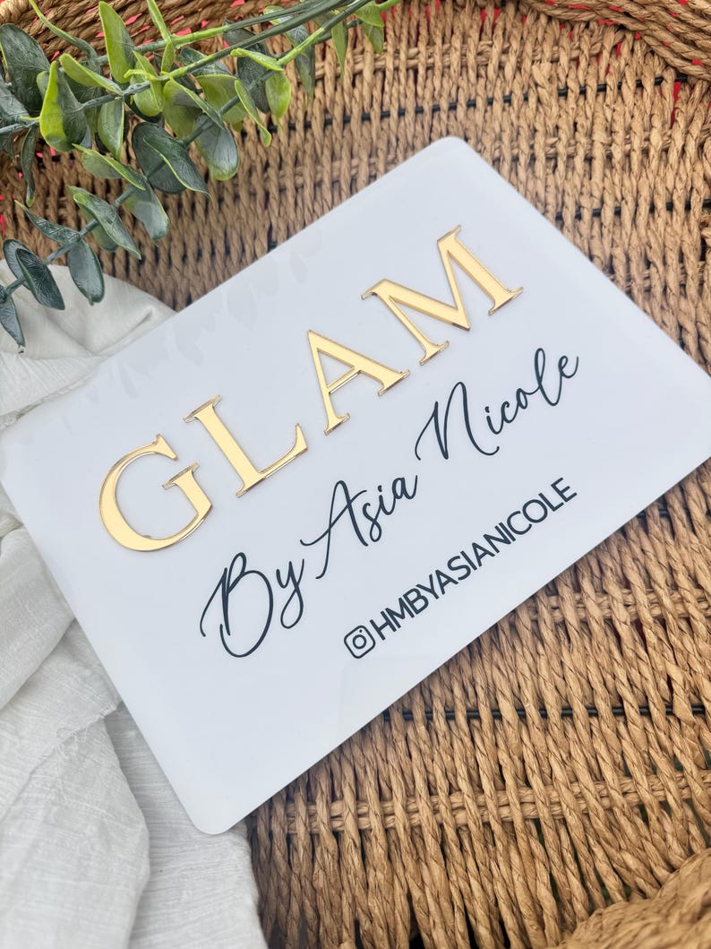 K&ouml;nnte beinhalten: Ein rechteckiges wei&szlig;es Acrylschild mit der goldenen erhabenen Aufschrift GLAM, dem schwarzen Schreibschrifttext By Asia Nicole und dem darunter gedruckten schwarzen Text @HMBYASIANICOLE. Das Schild hat abgerundete Ecken und eine glatte Oberfl&auml;che.