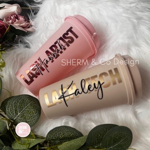 May include: Two pink and beige reusable coffee cups with the text "Lash Artist" and "Lash Tech" in gold and black lettering. The cups are on a white fluffy surface.