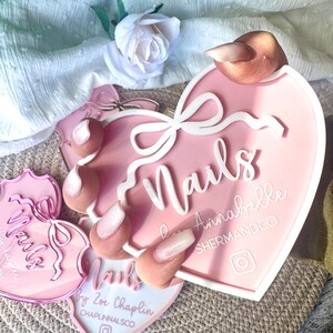 May include: Pink heart-shaped nail art display with the text "Nails" in white cursive lettering. The display also features a white bow and the text "Annabelle ShermanDCO" in white lettering. The display is surrounded by a white background.