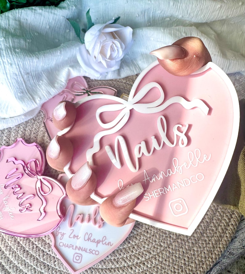 May include: Pink heart-shaped nail art display with the text "Nails" in white cursive lettering. The display also features a white bow and the text "Annabelle ShermanDCO" in white lettering. The display is surrounded by a white background.
