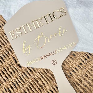 May include: A beige hand mirror with gold lettering that reads "ESTHETICS by Brooke". The mirror also features the Instagram handle "@BROOKEHALLESTHETICS" and an Instagram logo. The mirror is set against a woven background.
