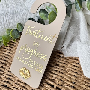 Puede incluir: Un colgador de puerta beige con la inscripción dorada "Treatment in progress PLEASE DO NOT DISTURB". Un diseño de labios dorados adorna la parte inferior. El colgador es de material liso.