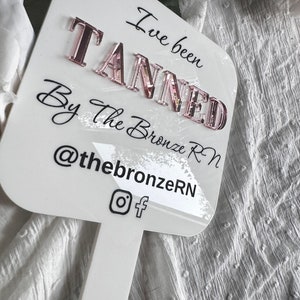 May include: White sign with black text that reads "I've been TANNED By the Bronze RN @thebronzerN" with Instagram and Facebook icons. The sign is attached to a white stick.