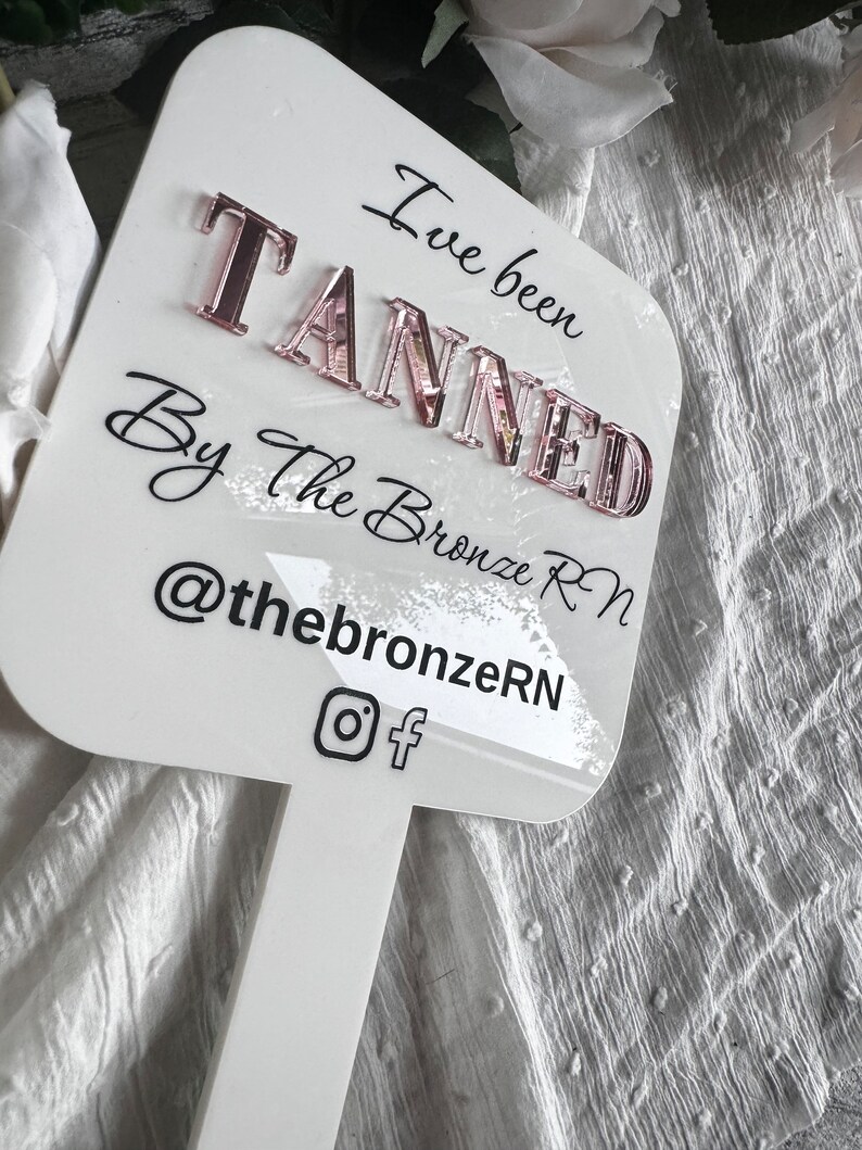 May include: White sign with black text that reads "I've been TANNED By the Bronze RN @thebronzerN" with Instagram and Facebook icons. The sign is attached to a white stick.