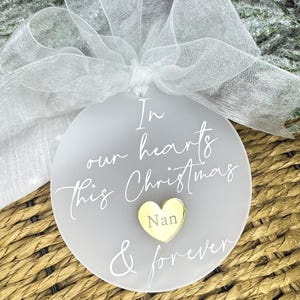 May include: A frosted glass Christmas bauble with the inscription "In our hearts this Christmas & forever." A gold heart with the word "Nan" is in the center. The bauble is tied with a sheer white ribbon.