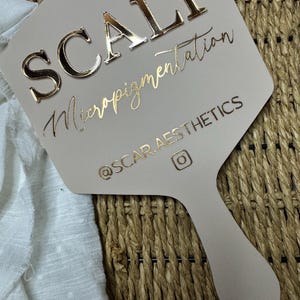 May include: A beige handheld mirror featuring the text SCALP Micropigmentation in raised gold lettering, with the handle and lower section displaying the social media handle @SCAR.AESTHETICS and an Instagram icon in gold foil.