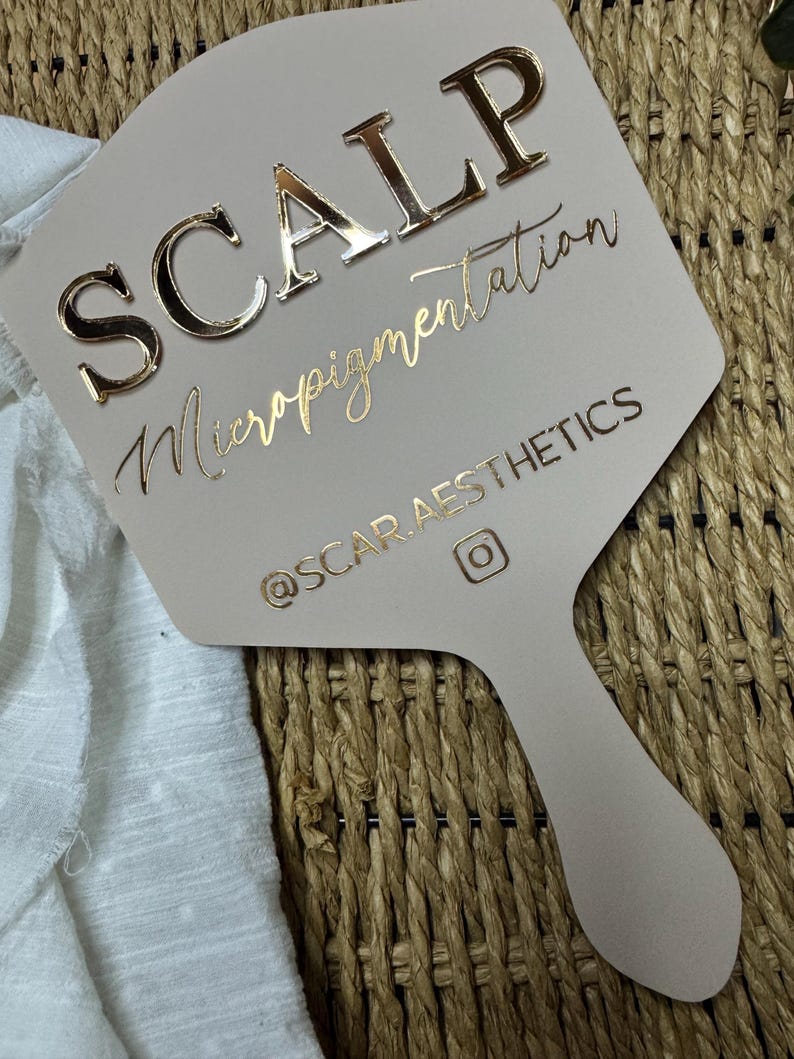 May include: A beige handheld mirror featuring the text SCALP Micropigmentation in raised gold lettering, with the handle and lower section displaying the social media handle @SCAR.AESTHETICS and an Instagram icon in gold foil.