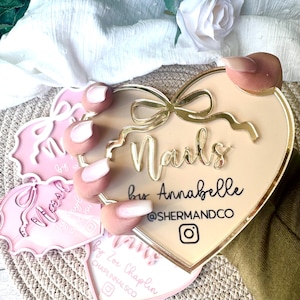 May include: A heart-shaped sign with a gold border and a gold bow. The sign says "Nails by Annabelle @shermandco" with an Instagram icon.