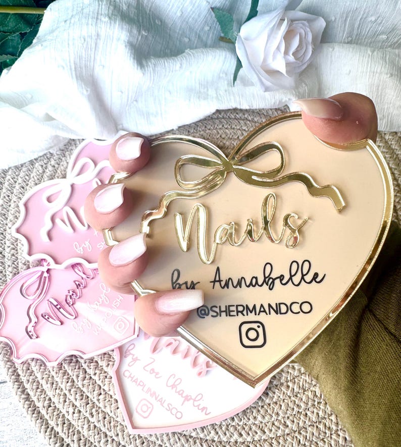 May include: A heart-shaped sign with a gold border and a gold bow. The sign says "Nails by Annabelle @shermandco" with an Instagram icon.