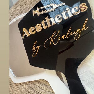 May include: Aesthetic hand mirror with a black surface and white handle. The mirror features the text "Aesthetics by Kiarleigh" in gold and silver lettering, along with a syringe graphic.