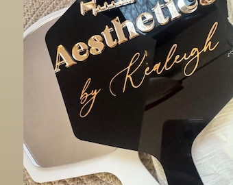 May include: Aesthetic hand mirror with a black surface and white handle. The mirror features the text "Aesthetics by Kiarleigh" in gold and silver lettering, along with a syringe graphic.