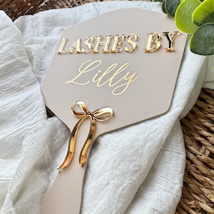 May include: A beige hand mirror with gold lettering that reads "LASHES BY Lilly". The mirror has a gold bow accent at the base of the handle. The mirror is resting on a white cloth and a woven basket.