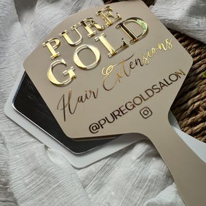 May include: A beige handheld mirror featuring the text PURE GOLD Hair Extensions and the social media handle @PUREGOLDSALON in gold lettering. The words PURE GOLD are raised in a metallic finish, while the remaining text is in a script font.