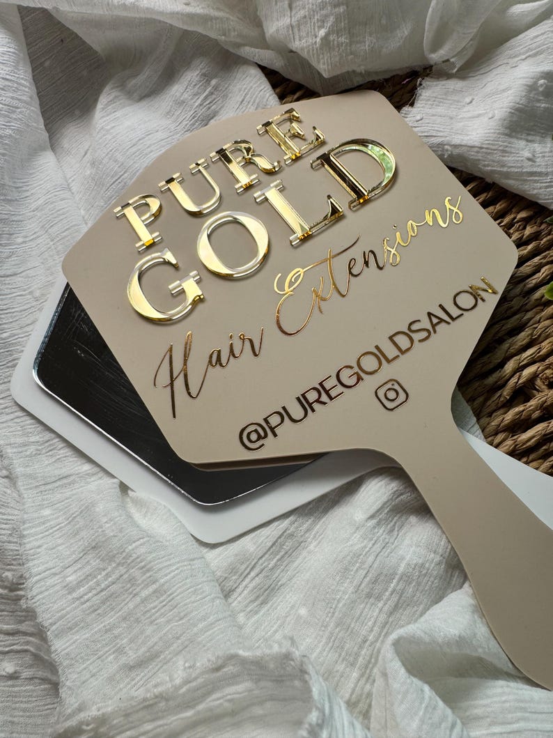 May include: A beige handheld mirror featuring the text PURE GOLD Hair Extensions and the social media handle @PUREGOLDSALON in gold lettering. The words PURE GOLD are raised in a metallic finish, while the remaining text is in a script font.
