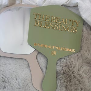 May include: A sage green handheld mirror with a handle and a gold-colored text overlay that reads THE BEAUTY BLESSINGS and @THEBEAUTYBLESSINGS with an Instagram icon. A second beige handheld mirror is partially visible underneath the green one.