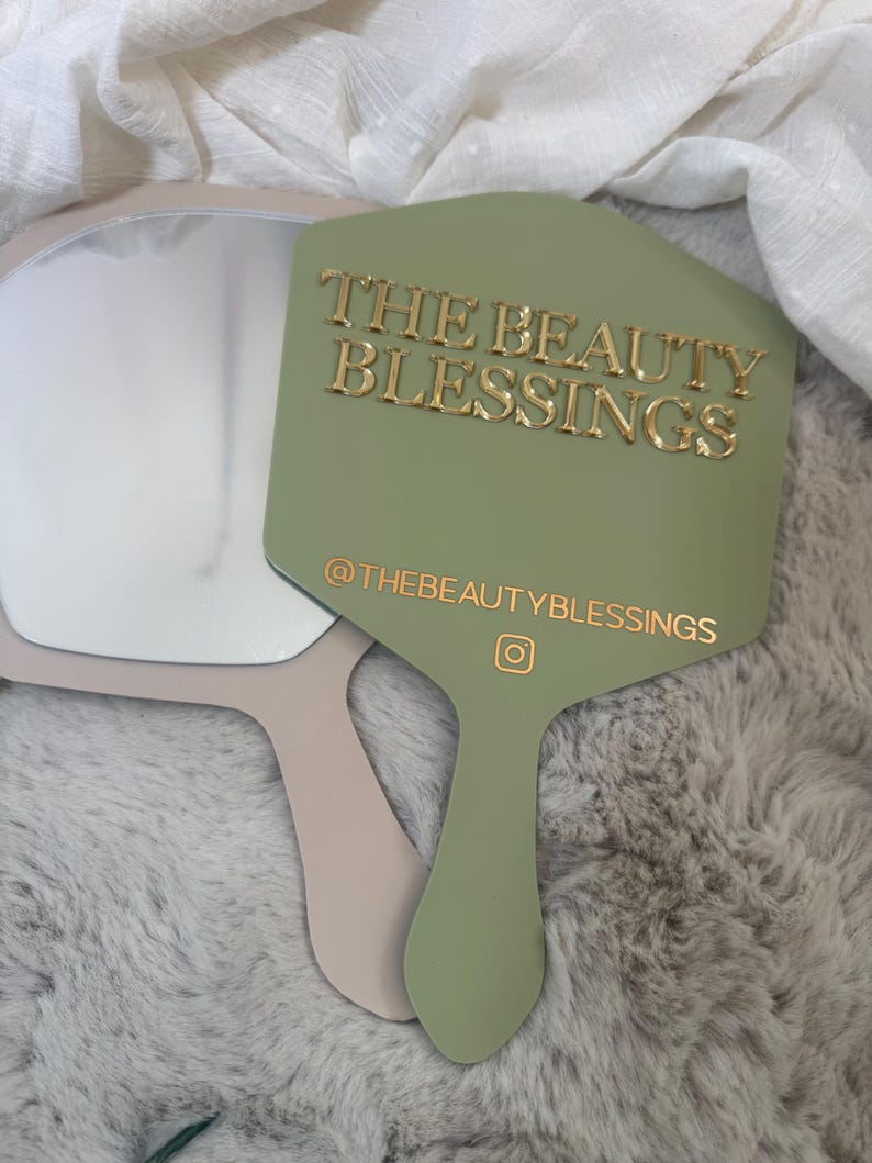 May include: A sage green handheld mirror with a handle and a gold-colored text overlay that reads THE BEAUTY BLESSINGS and @THEBEAUTYBLESSINGS with an Instagram icon. A second beige handheld mirror is partially visible underneath the green one.