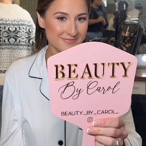 May include: A pink hand mirror with gold lettering that reads "BEAUTY By Carol". The mirror also has the Instagram handle "BEAUTY_BY_CAROL_". The person in the image is wearing a white shirt.
