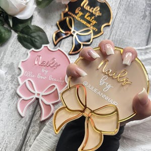 Pode incluir: Três suportes redondos de acrílico para arte de unhas com laços dourados. Os suportes são preto, rosa e bege. O suporte preto diz "Nails by Katy" e o suporte rosa diz "Nails by Ollie Bow Beauty". O suporte bege diz "Nails by" e tem o nome de usuário do Instagram "@JULIEANNHAIR" escrito abaixo.