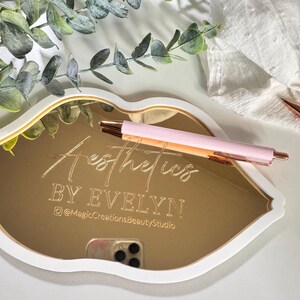 May include: Gold mirror tray shaped like a lip with the text "Aesthetics by Evelyn @MagicCreationsBeautyStudio" engraved on the surface.