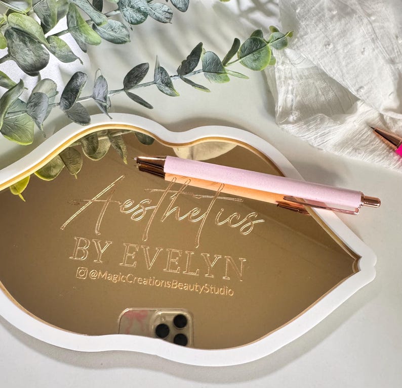 May include: Gold mirror tray shaped like a lip with the text "Aesthetics by Evelyn @MagicCreationsBeautyStudio" engraved on the surface.