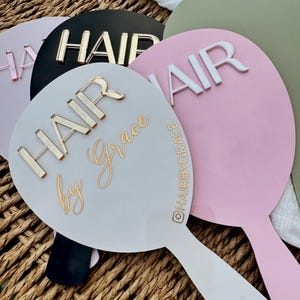 Puede incluir: Una colección de espejos de mano redondos en varios colores, incluyendo rosa, negro y blanco. Cada espejo presenta la palabra "HAIR" en letras en relieve. Algunos espejos tienen texto adicional, como "Hair by Grace" y un identificador de Instagram.