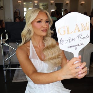 May include: A white hexagonal handheld mirror with the text GLAM by Asia Nicole and HMBYASIANICOLE printed in gold and black. The mirror is held by a person with long blonde wavy hair wearing a white corset-style top with lace trim details.