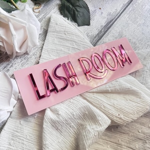 Puede incluir: Un letrero de acrílico rosa con el texto "LASH ROOM" en letras rosas reflectantes. El letrero está sobre un fondo de tela blanca.