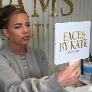 May include: A white rectangular handheld mirror featuring the gold text FACES BY KATE and the social media handle @facesby_kate. The mirror is held by a person wearing a grey long-sleeve button-up sweatshirt with a ribbed collar and cuffs.