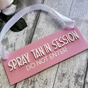Op de afbeelding: Een roze bord met witte tekst die "Spray Tan in Session Do Not Enter" zegt. Het bord heeft een wit lint dat aan de bovenkant is bevestigd om het op te hangen.