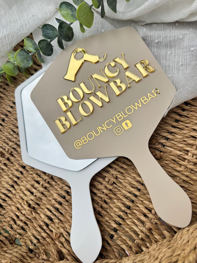 May include: A beige handheld mirror with a hexagonal shape featuring a gold acrylic hair dryer icon and the text BOUNCY BLOWBAR. Below the text is the handle @BOUNCYBLOWBAR with Instagram and Facebook icons in gold lettering.