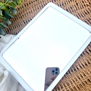 K&ouml;nnte beinhalten: Ein rechteckiger Spiegel mit einem glatten wei&szlig;en Rahmen und abgerundeten Ecken. Die reflektierende Glasoberfl&auml;che zeigt das klare Spiegelbild eines Smartphones mit einem Multilinsen-Kamerasystem, das in einer Hand gehalten wird.