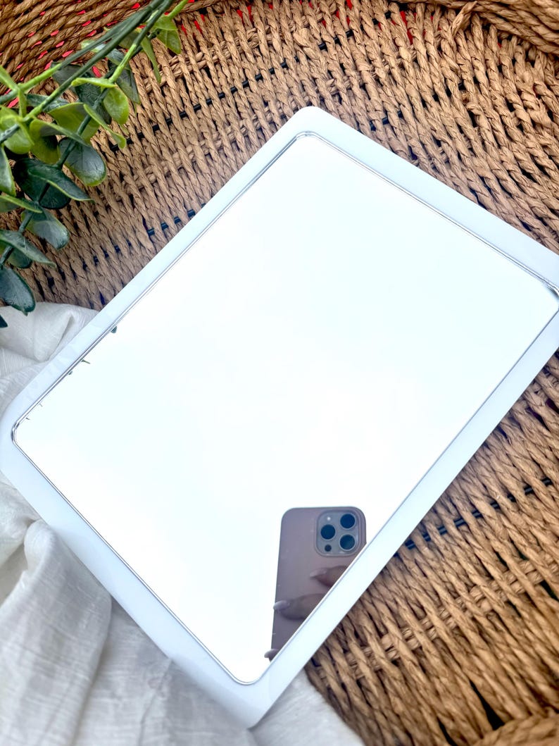 K&ouml;nnte beinhalten: Ein rechteckiger Spiegel mit einem glatten wei&szlig;en Rahmen und abgerundeten Ecken. Die reflektierende Glasoberfl&auml;che zeigt das klare Spiegelbild eines Smartphones mit einem Multilinsen-Kamerasystem, das in einer Hand gehalten wird.
