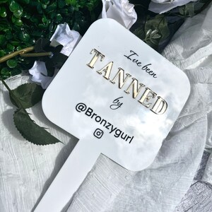 May include: White sign with gold lettering that says "I've been tanned by @Bronzygurl" with an Instagram icon. The sign is attached to a white stick.
