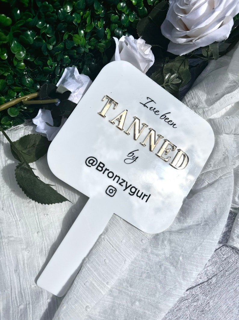 May include: White sign with gold lettering that says "I've been tanned by @Bronzygurl" with an Instagram icon. The sign is attached to a white stick.