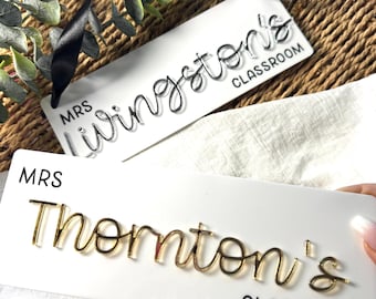 May include: Two white rectangular classroom signs. One reads "MRS Livingston's CLASSROOM" with silver lettering and a black ribbon. The other says "MRS Thornton's CLASSROOM" with gold lettering.