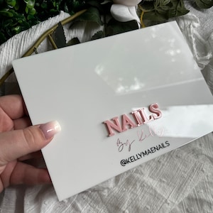 May include: White acrylic sign with the text "NAILS by Kelly" in pink letters and the text "@KELLYMAENAILS" in black letters.