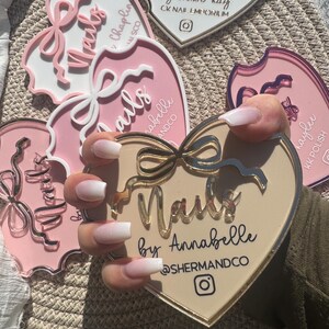 May include: Close-up of a hand with white ombre nails holding a heart-shaped sign that reads "Nails by Annabelle @SHERMANDCO". The sign is beige with gold accents and a pink bow.