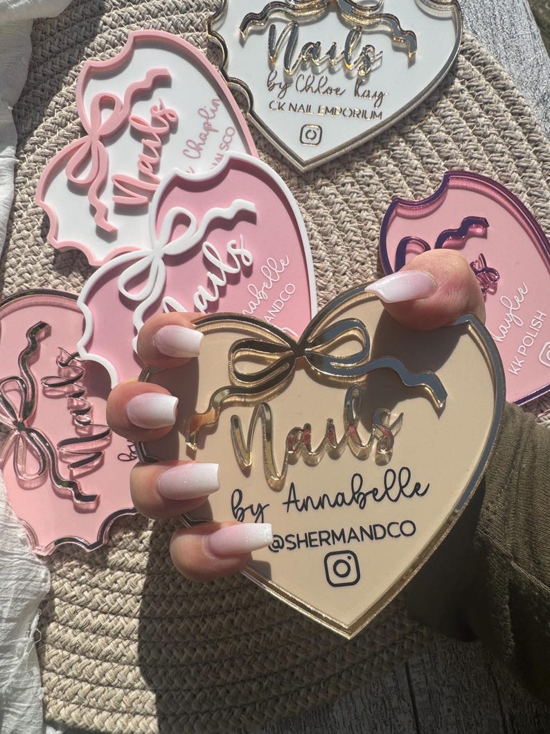 May include: Close-up of a hand with white ombre nails holding a heart-shaped sign that reads "Nails by Annabelle @SHERMANDCO". The sign is beige with gold accents and a pink bow.