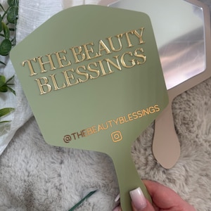 May include: A sage green handheld mirror with a hexagonal shape. The front features the gold text THE BEAUTY BLESSINGS and the Instagram handle @THEBEAUTYBLESSINGS with an icon. A second, plain beige mirror is partially visible behind it.