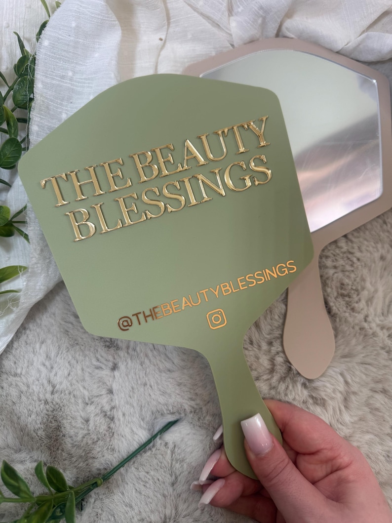 May include: A sage green handheld mirror with a hexagonal shape. The front features the gold text THE BEAUTY BLESSINGS and the Instagram handle @THEBEAUTYBLESSINGS with an icon. A second, plain beige mirror is partially visible behind it.